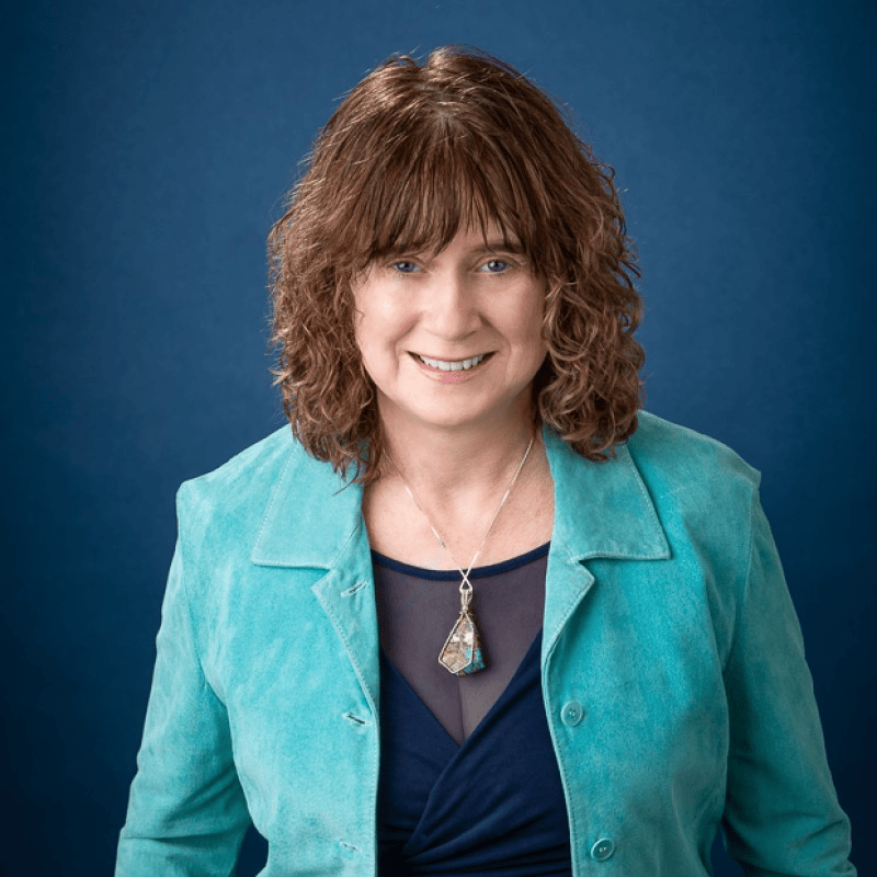 Professional woman smiling in a teal jacket against a blue background, representing expertise in behavioral change for AI adoption.