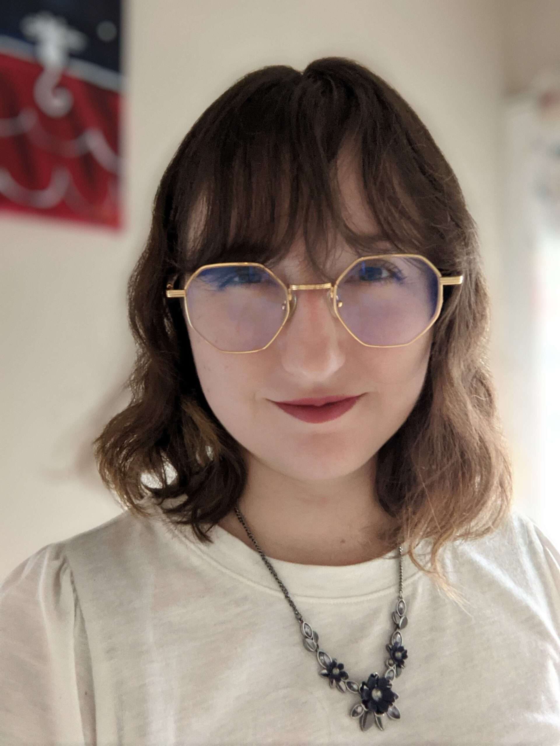Young woman with glasses and a floral necklace, smiling in a bright indoor setting.